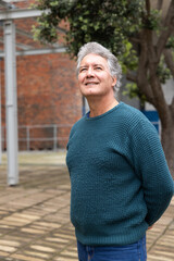 Fototapeta premium Senior male standing in paved courtyard wearing teal knit sweater and blue jeans, looking upward