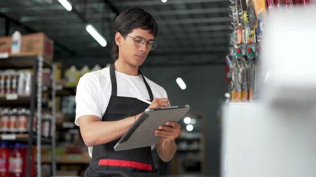 Young male employee checking stock and taking notes on a digital tablet in a hardware store aisle