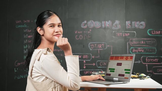 Smart girl using laptop coding or programming system at blackboard. Happy high school student working on laptop while turnaround and showing miniheart and looking at camera at STEM class. Edification