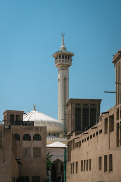 Mosque minaret and white dome rising above traditional wind towers and coral-stone walls in the historic district of Dubai, United Arab Emirates, under a clear blue sky