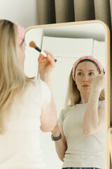 Naklejka premium Young woman applying makeup in front of a mirror at home. Natural beauty routine with soft light, skincare concept, and everyday self care lifestyle.
