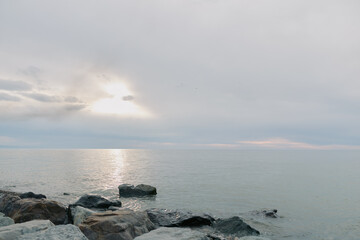 Fototapeta premium Calm sea with gentle waves and rocks in foreground under cloudy sky at sunrise. Peaceful coastal landscape with soft natural light reflecting on water surface.