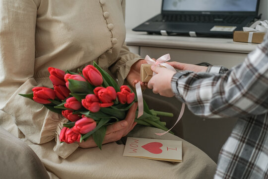 Close-up of mother holding red tulips and "I Love Mom" card while receiving a gift from her child. Warm domestic scene ideal for Mother's Day, remote work balance and spring celebrations