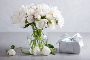 Still life with a bouquet of white peonies in a glass vase, a silver gift box, tied with a satin...