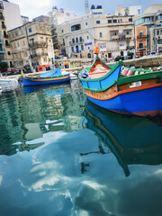 Fototapeta premium Colorful boats floating in a Maltese harbor near old waterfront buildings. The image conveys the concept of coastal culture and traditional Mediterranean scenery.