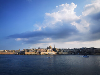 Naklejka premium Malta skyline rising above calm blue water beneath dramatic cloud formations. The image conveys the concept of coastal landscape and open Mediterranean scenery.