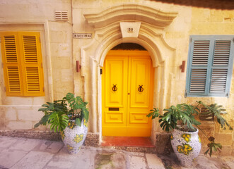 Fototapeta premium Yellow wooden door on a Maltese stone facade framed by decorative plants. The image conveys the concept of architectural detail and colorful Mediterranean style.