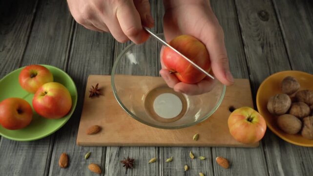 Hands slicing an apple over a glass bowl on a wooden cutting board, surrounded by fresh apples and assorted nuts in colorful bowls
