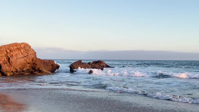 Waves washing up to sandy beach with a few large rocks in the water in warm glow of the setting sun