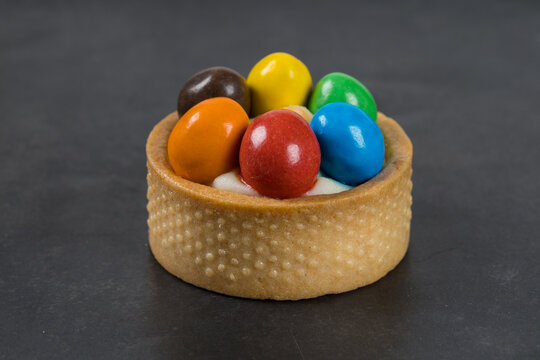 Small Round tartlet with vanilla custard and colorful dragees in the shape of Easter eggs. Happy Easter. Dark gray background. Close-up.