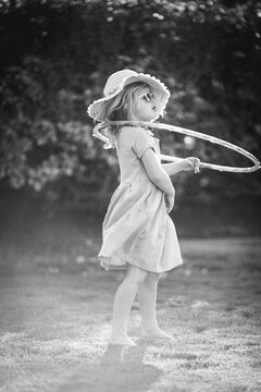 Monochrome imge of a child hula hooping in the backyard. It's a warm day and she's wearing a dress, sunglasses, and a sunhat.