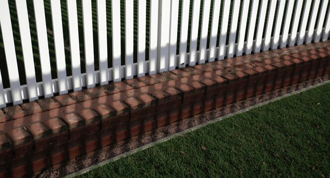 silhouettes cast by a textured red brick boundary and synthetic turf surrounded by white picket fence