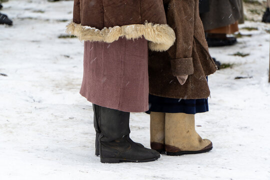 Lower body view showing women wearing traditional wool skirts heavy fur trimmed coats black leather boots and felt valenki standing on snow during winter historical event.
