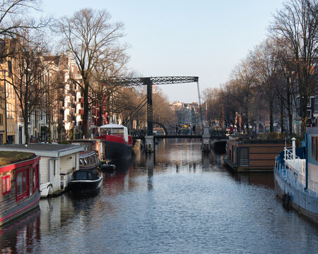 Historic Amsterdam Drawbridge and Houseboats - Iconic view of a wide canal in Amsterdam featuring a traditional metal drawbridge, residential houseboats, and Dutch architecture under a clear sky.