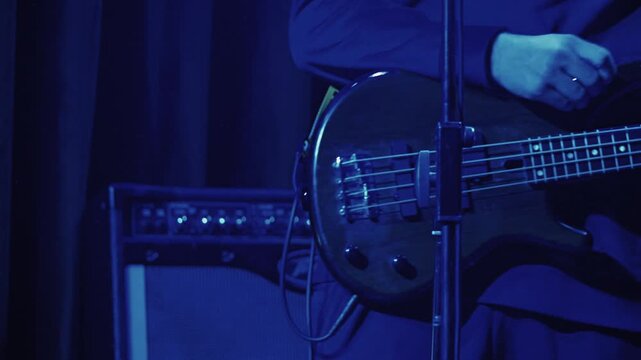 Close up of a bassist playing a red bass guitar on a dark stage. The musician's hands pluck the strings with an amplifier in the background. Moody lighting creates an energetic, live music concept