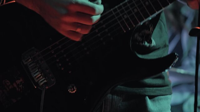 A close up a musician's hand strumming an electric guitar during a live performance. The energetic scene is lit by dramatic purple and red lights, creating a moody and intense atmosphere