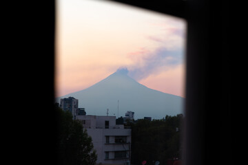 Popocatepetl volcano emitting smoke against pastel sunset sky viewed through dark window frame from interior space overlooking urban buildings in Puebla, Mexico © simonmayer