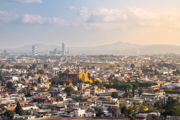 Naklejka premium Panoramic view of historic yellow Parish of San Andres Apostol in Cholula with modern Puebla skyline featuring Bicentenario Tower under hazy afternoon sky in Cholula, Puebla, Mexico