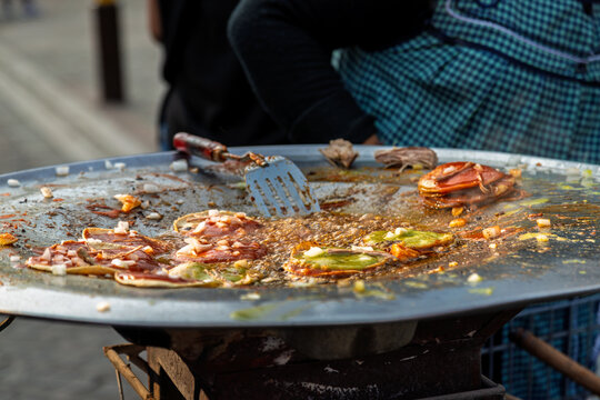 Close-up of traditional chalupas poblanas frying in bubbling oil on large metal comal with red and green sauces, onions and shredded meat at street food stall in Cholula, Puebla, Mexico