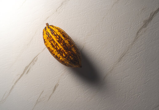 Mazorca de cacao entera sobre superficie de m&aacute;rmol blanco con iluminaci&oacute;n dram&aacute;tica