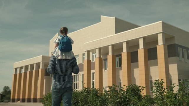 dad carries a little daughter on his shoulders to school, a girl with a school bag on his shoulders, walk around the schoolyard, see off to first grade, preschool education, father day, happy family.