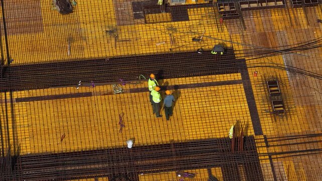 Aerial drone shot looking straight down at workers assembling dense steel rebar grid on formwork deck, highlighting structural reinforcement, teamwork, and building progress.