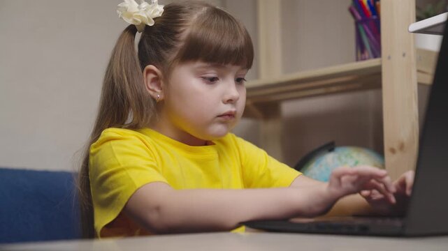 Small kid writes by pressing keyboard keys on computer. kid girl does her homework sitting at her desk. Home learning online laptop. student children on home e-learning. concept of American learning