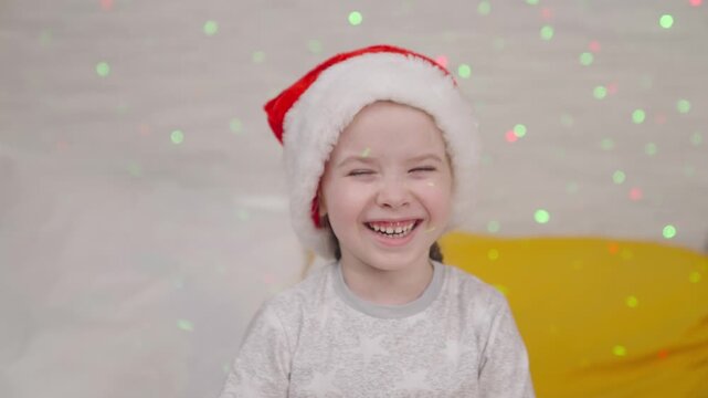 little child laughs in Santa hat, kid smile Christmas holidays, happy family, baby has fun playing bright colors of garland, festive mood in new year, waiting for miracle child dream, time for gifts.