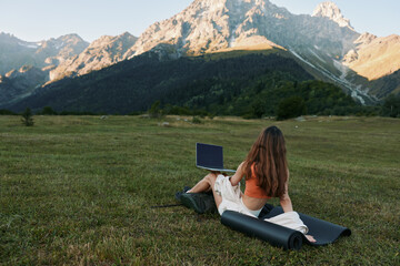 Naklejka premium A person sits on a grassy meadow with a laptop, distant mountains in the background, creating a peaceful outdoor work moment and a sense of calm focus for creative tasks and learning.