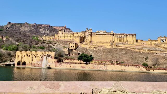 A bright daytime  showing the historic Amer Fort and surrounding defensive walls spread across the Aravalli hills, with Maota Lake in the foreground.