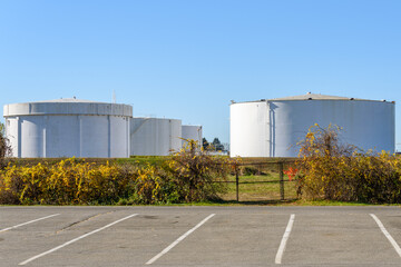 Obraz premium Rows of large white steel fuel tanks in an oil terminal under blue clear sky in autumn. A deserted car park is in foreground.
