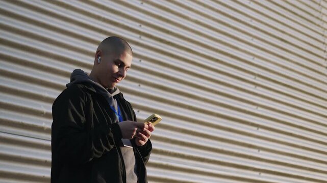 Young bald man using smartphone and listening to music with wireless earbuds against a corrugated wall outdoors in the sunlight