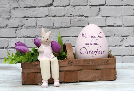 Oster-Dekoidee: Rosa Osterei mit der Aufschrift, wir w&uuml;nschen ein frohes Osterfest mit lila Tulpen und einen Dekohasen im Spankorb.