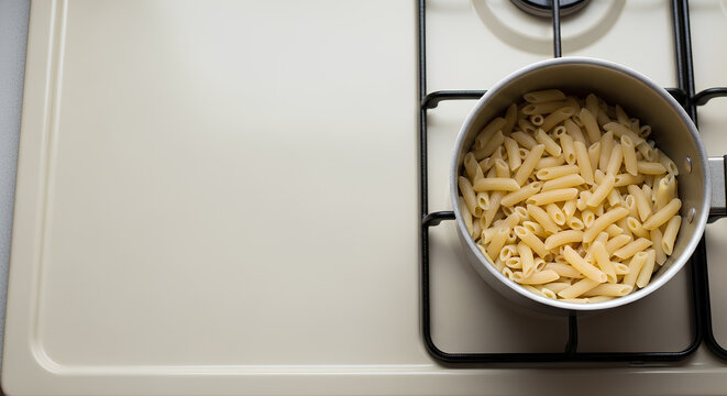 Pot with a small amount of slightly overcooked plain pasta sitting on the right third of a pale beige stove surface, large empty left area serving as negative space.