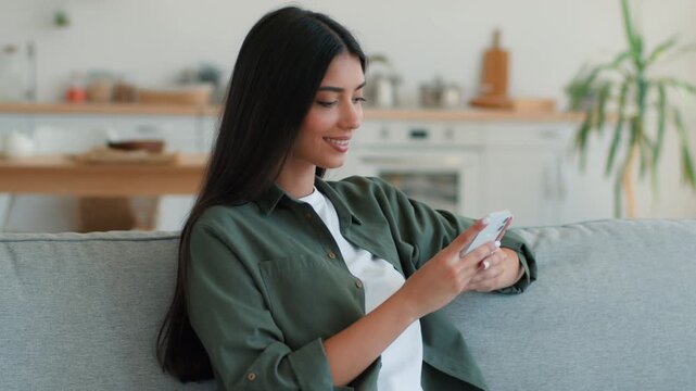 Happy smiling Indian woman using mobile phone at home apartment looking at camera satisfied chatting shopping online message app Arabian girl female holding smartphone internet communication indoors