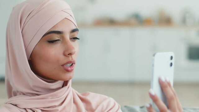 Smiling friendly Arabian Muslim woman in hijab talking video call indoors at home female Islamic girl young lady using mobile phone vlog blog smartphone chatting conference online virtual meeting