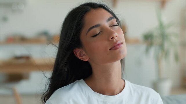 Calm Indian Arabian woman enjoying cool air breeze closed eyes at home indoors female relaxing feeling fresh wind blowing hair conditioner cooling fan comfort climate control system in house apartment