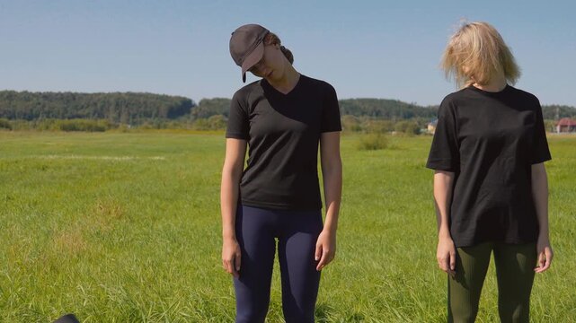 Two young athletic women standing in a sunny green field, performing neck rotation and stretching exercises as a warm-up before a workout, promoting a healthy and active lifestyle