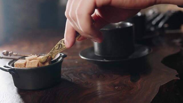 Closeup of male caucasian hand picking brown sugar cube, delicate brass tongs lifting brown sugar from vintage bowl and add into tea in black ceramic cup on wooden table. Old loft and stylish vibe