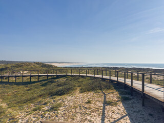 Fototapeta premium Coastal wooden walkway winding through natural sand dunes towards the Atlantic Ocean under a bright blue sky at Furadouro beach in Ovar Portugal