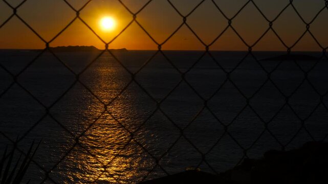 Golden sunset over ocean seen through wire mesh fence. Warm sunlight reflects on the calm ocean as the sun sets behind small islands, creating a picturesque scene viewed through a wire mesh fence
