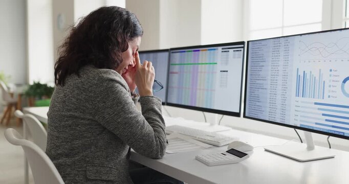 Woman accountant in office shows stress and headache. She works at computer screens reviewing spreadsheets, feeling fatigue and eye strain from long hours. Theme of burnout at modern workplace.