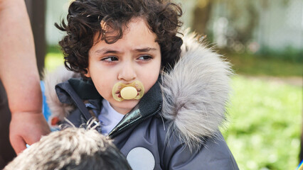 Toddler with curly hair wearing a parka and pacifier outdoors, looking away © Довидович Михаил