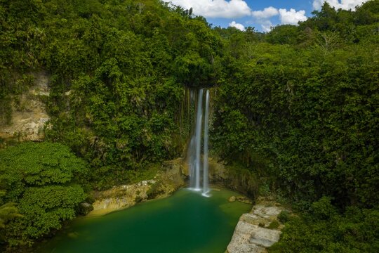 Dramatic aerial view of Camugao Falls in Balilihan, Bohol. Tall waterfall plunging into a deep pool within a massive limestone cliff amphitheater and lush forest.