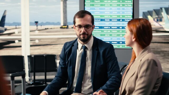 Colleagues on business trip seated together in busy airport waiting area, delegation talking over briefing notes. Man and woman in business attire for corporate travel, waiting for boarding.
