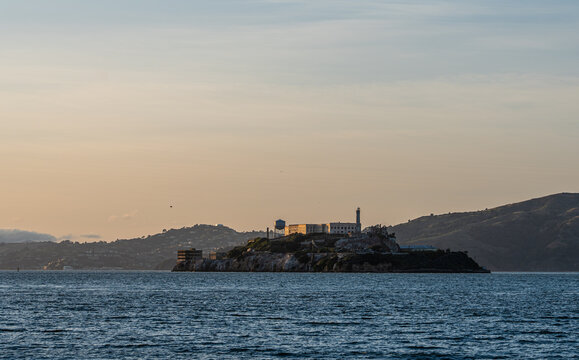 The historic Alcatraz Island prison complex sits isolated amidst the shimmering waters of the San Francisco Bay under a soft, atmospheric sky.