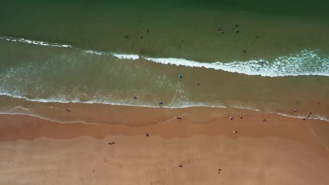 This aerial view shows beachgoers enjoying the shoreline near Caparica, Portugal. People swim, sunbathe, and relax on the sand while waves roll in.