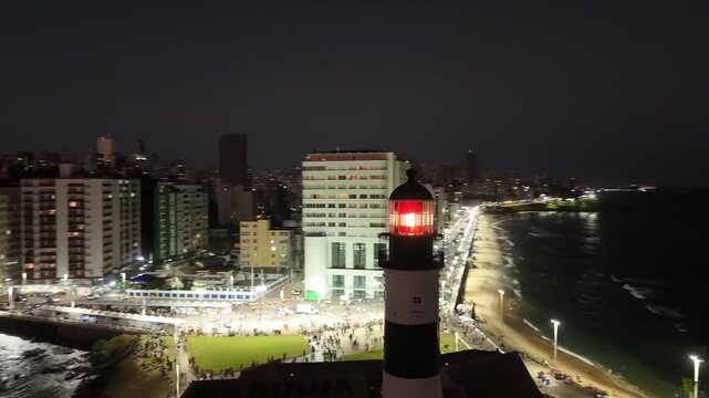 Close aerial view of Farol da Barra lighthouse beacon glowing red with illuminated city buildings and coastline at night in the background