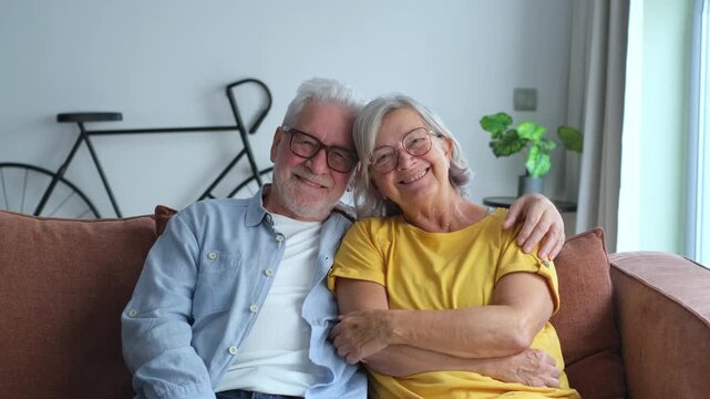 Joyful senior couple sitting embraced on a comfortable sofa, smiling warmly at the camera, then sharing an affectionate glance, embodying enduring love and companionship.