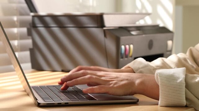 Woman using printer while working on laptop at wooden desk indoors, closeup